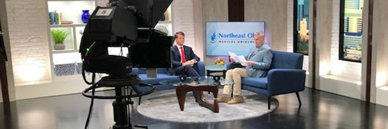 two men seated in a TV studio. Large camera in the foreground