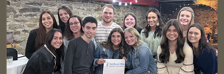 A group of smiling pharmacy students holding a NEOMED sign.