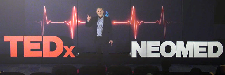 A man gestures while standing on a stage, speaking.